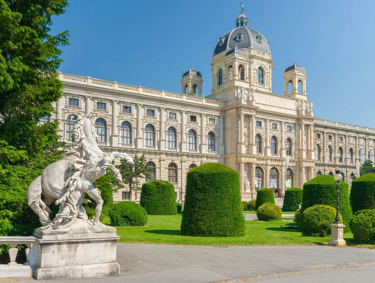 A historic building with elaborate architecture, surrounded by manicured bushes and a statue of a horse and rider in the foreground.