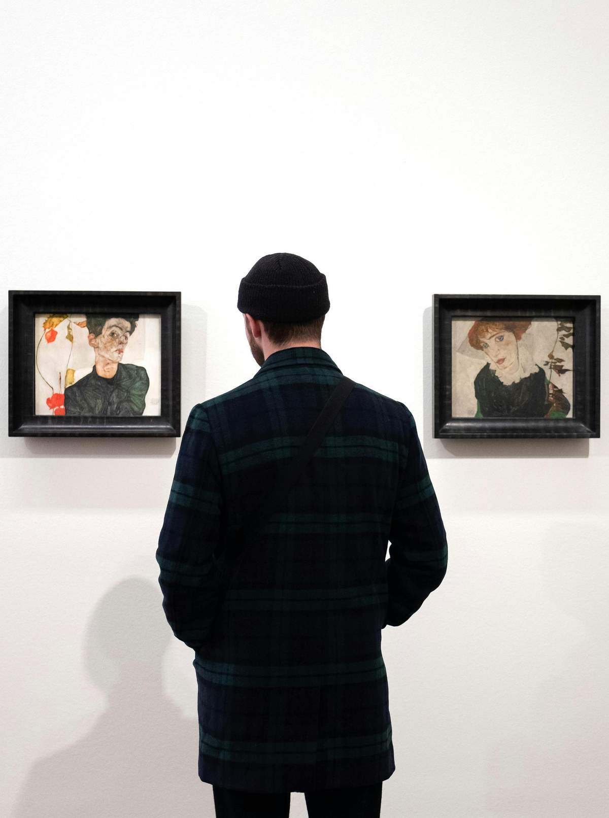 Leopold Museum: Fast Track Ticket A person wearing a black beanie and dark plaid jacket stands in front of two framed artworks on a white wall.