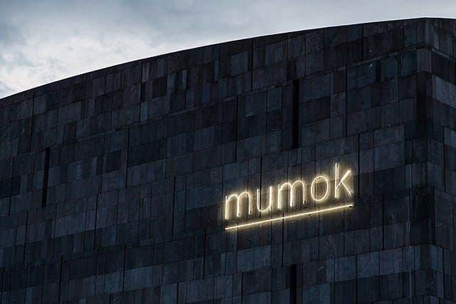 mumok: Museum of Modern Art Ludwig Foundation: Skip The Line A dark, modern building facade with the illuminated word "mumok" in neon lights against a cloudy sky.