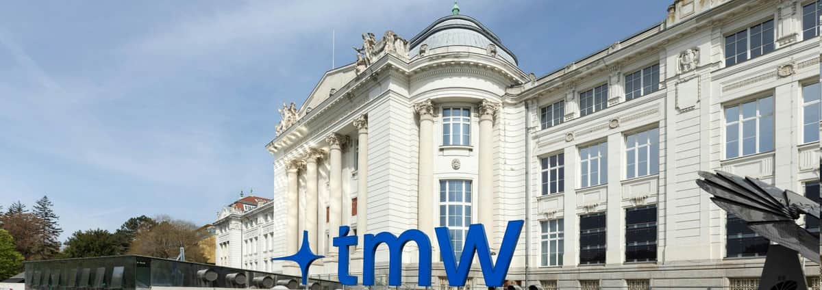 Vienna Technical Museum: Skip The Line Ticket Ornate white building with tall columns and sculptures above the entrance, large blue 'tmW' sign in the foreground.