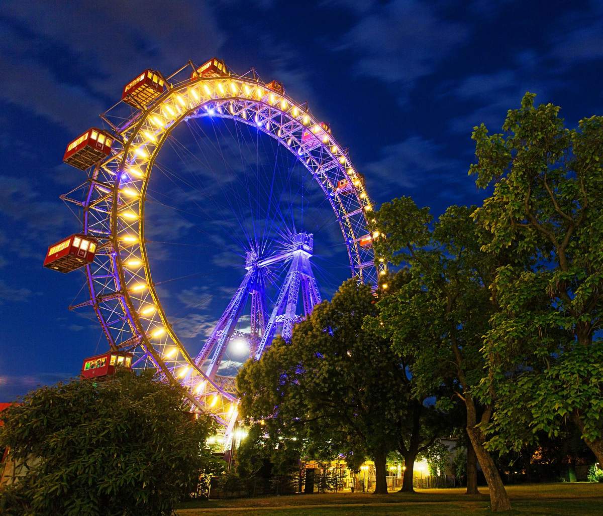 Vienna's Giant Ferris Wheel: Skip The Line Illuminated Ferris wheel with red cabins at night, surrounded by trees, against a cloudy sky.