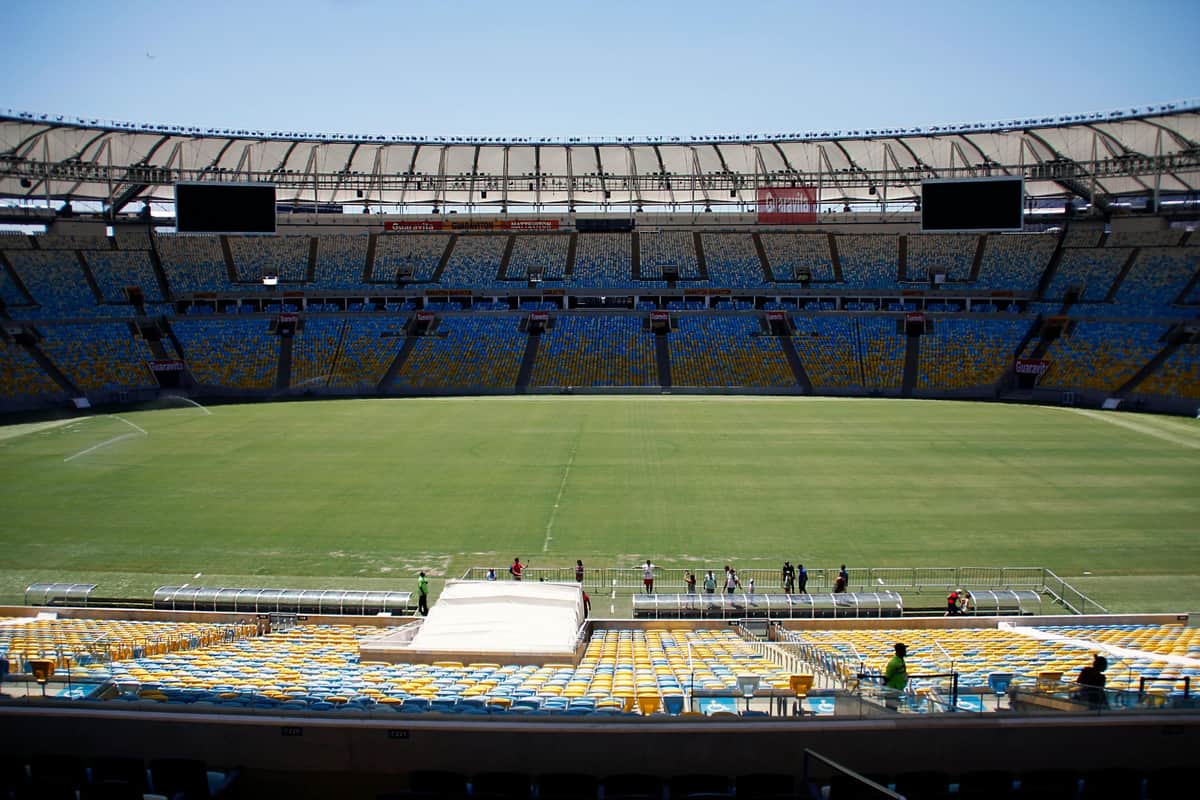 Empty stadium with multi-colored seats, green field, and a few people standing near the sideline under a clear sky.