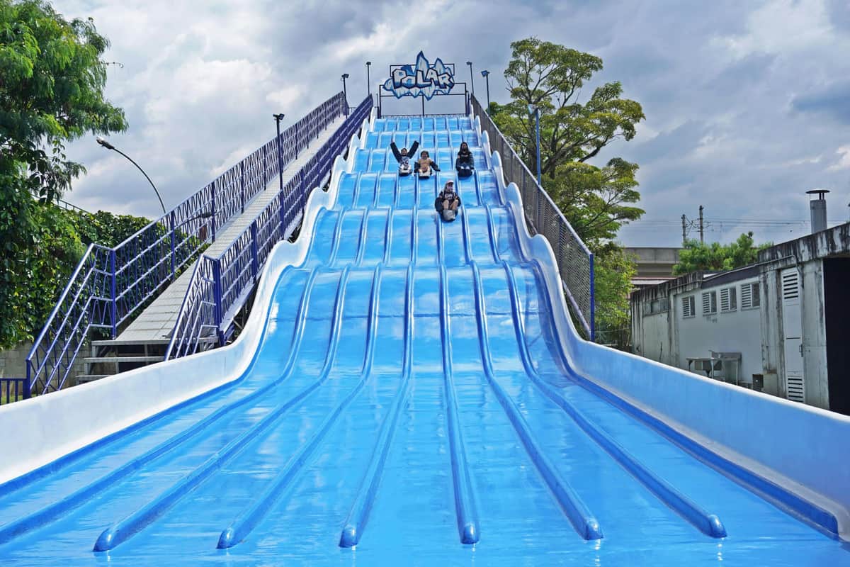 Three people slide down a large, multi-lane blue slide labeled "Polar" under a cloudy sky. Trees and a walkway are visible nearby.