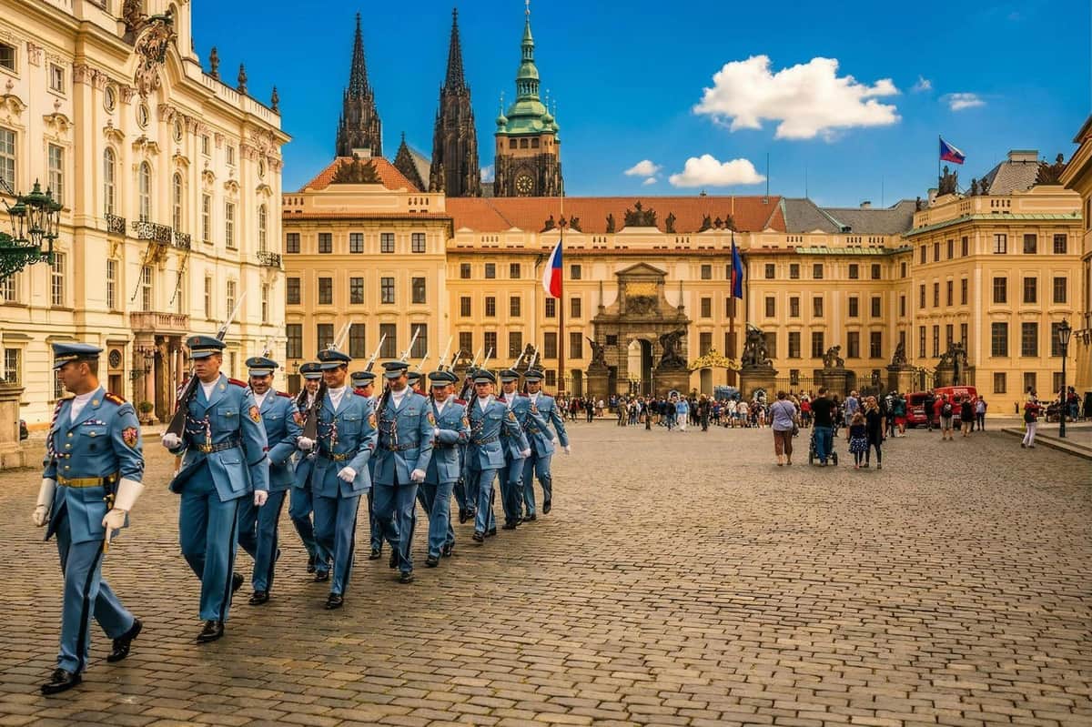 Prague: Hradčany Guided Tour + Fast Track Castle Entry + Audio Guide A procession of uniformed guards marching in a historic square with ornate buildings and a cathedral in the background.