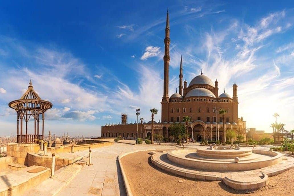 A grand mosque with multiple domes and minarets under a bright blue sky with scattered clouds, surrounded by a courtyard.