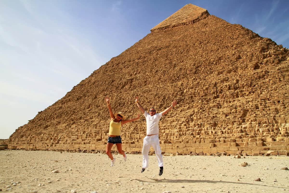 Two people jumping in front of the Great Pyramid of Giza, clear sky in the background.