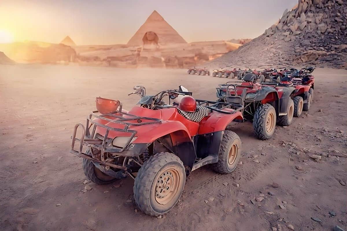 Several red all-terrain vehicles parked on a desert landscape with pyramids and rocky surroundings in the background.