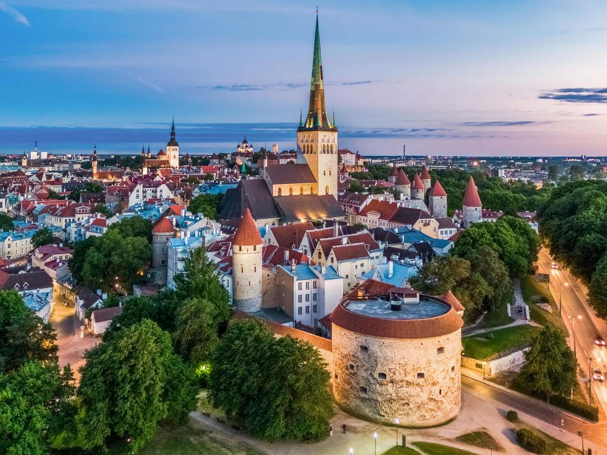 Aerial view of a medieval European city with numerous red-roofed buildings, a large church spire, and surrounding greenery at dusk.