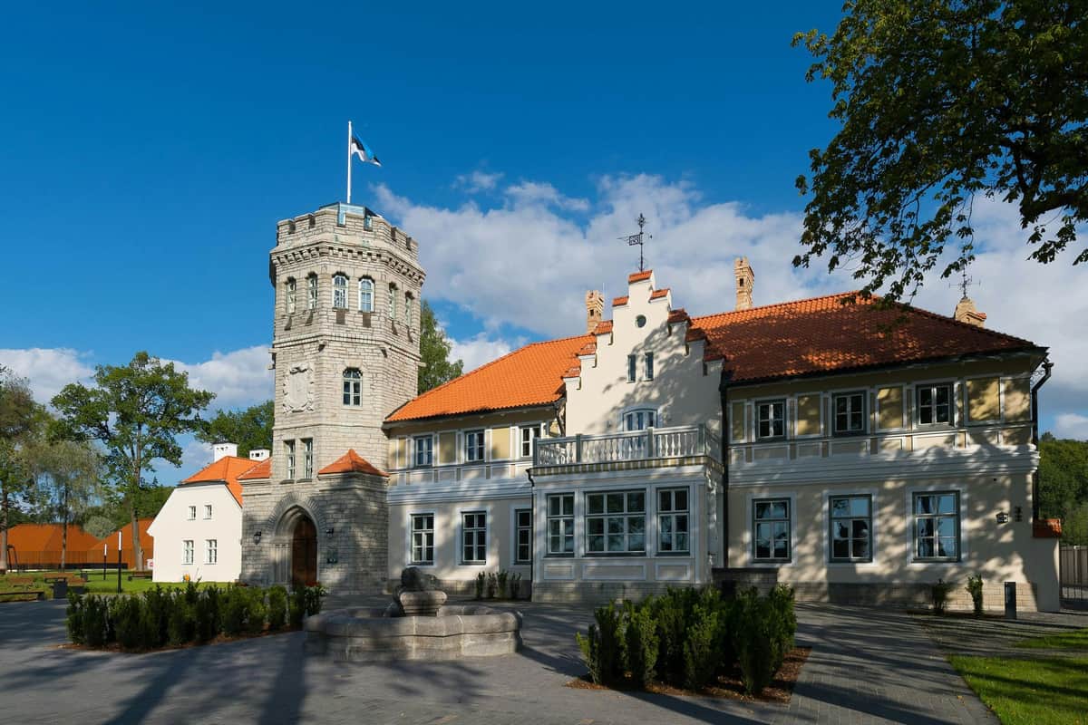 Maarjamäe History Center: Skip The Line Ticket Historic building with a stone tower and an orange-tiled roof beneath a blue sky, featuring a flag atop the tower and well-kept shrubs.