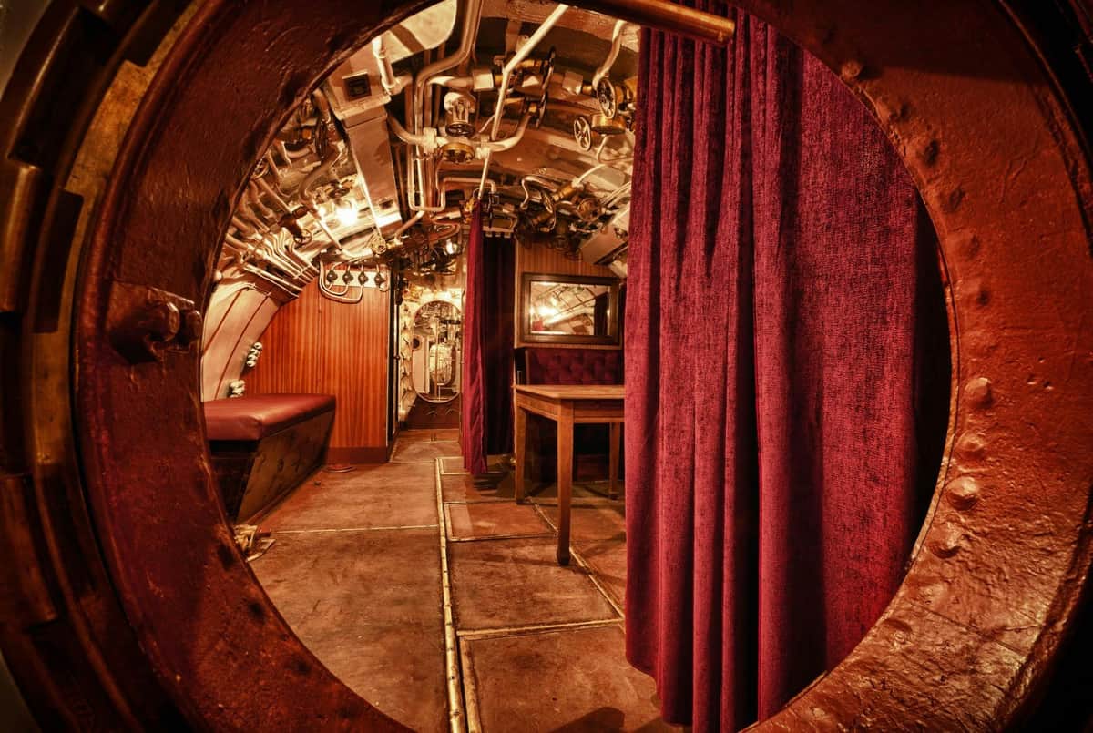 View into a submarine interior with red curtains, metal pipes, a wooden table, and a bench.