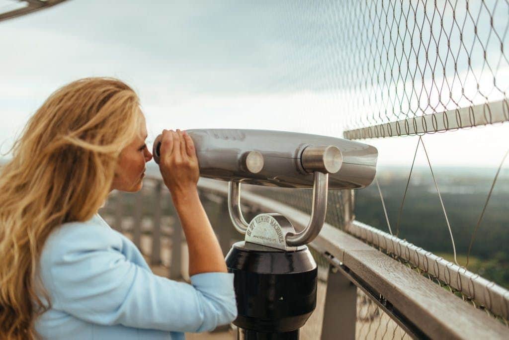 Tallinn TV Tower: Skip The Line Ticket A person looking through a coin-operated viewer at an observation deck, with a safety fence and blurry landscape in the background.