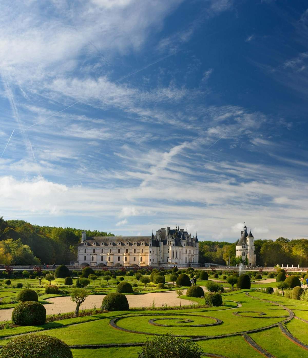 A picturesque castle with landscaped gardens under a blue sky with scattered clouds and trees in the background.