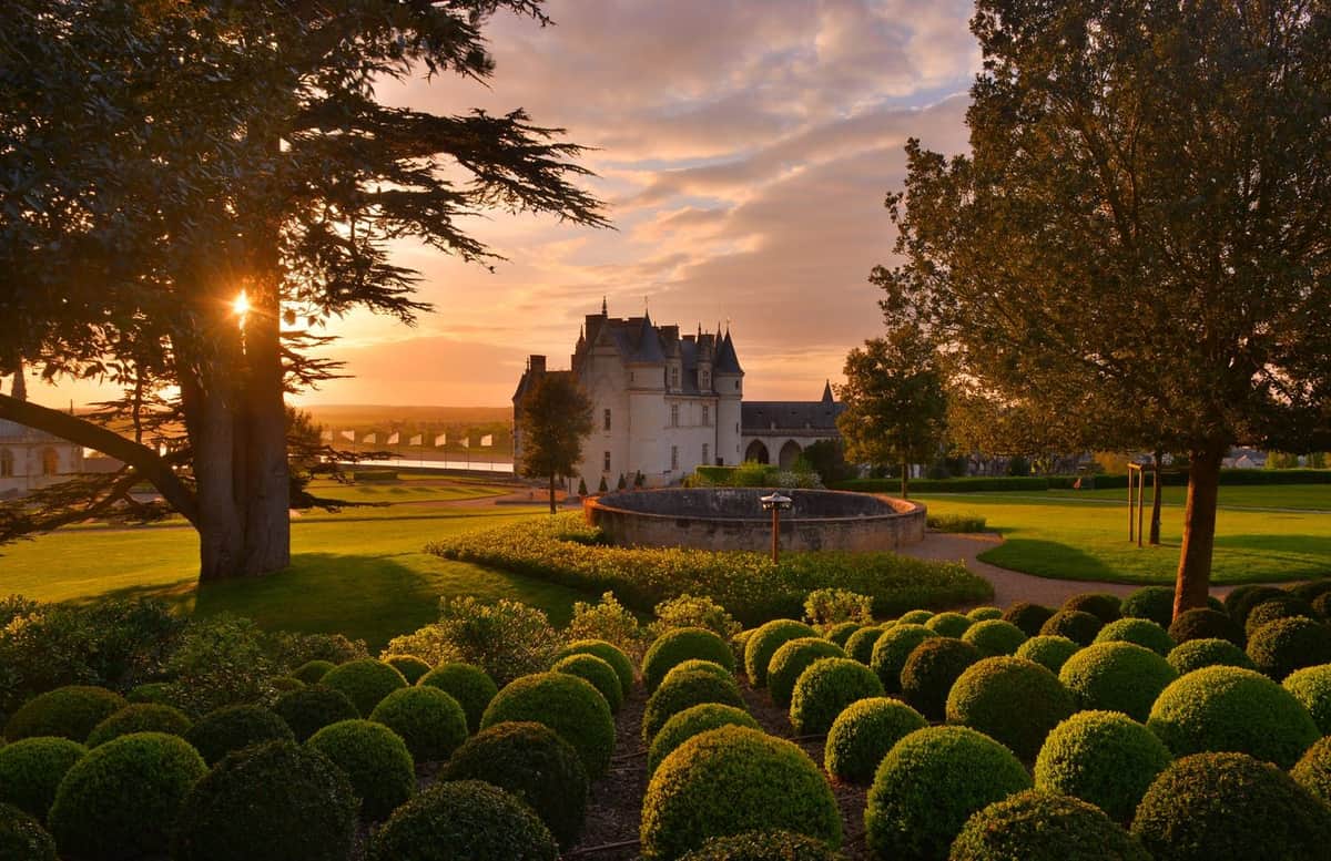 Château Royal d'Amboise + Château du Clos Lucé Manicured garden with round hedges, trees, and a stone building in the background at sunset.