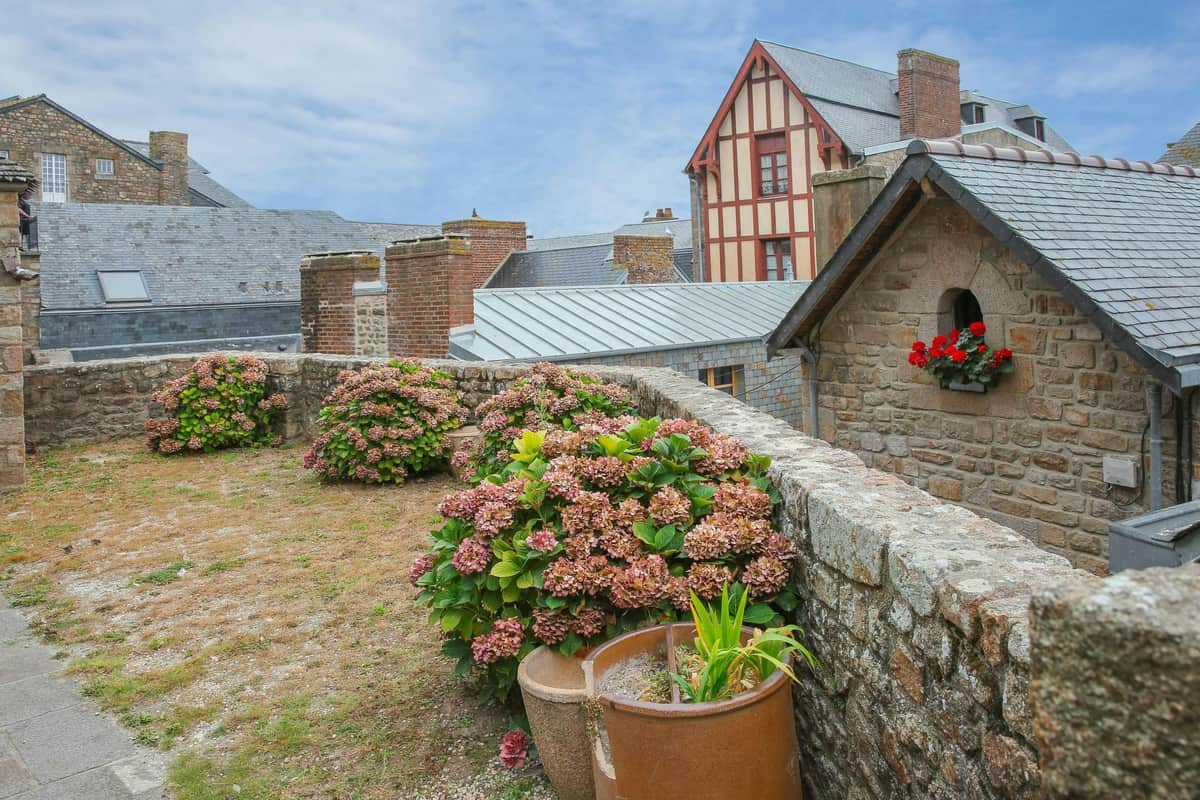 Mont Saint-Michel: Entry Ticket and Guided Tour + Transport from Paris Rooftop garden with blooming hydrangeas, stone walls, and a red-trimmed building in the background under a partly cloudy sky.