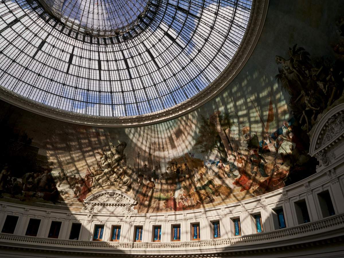 Bourse de Commerce - Pinault Collection: Priority Access Ticket Glass-domed ceiling with intricate iron framework, casting shadows on a fresco-adorned wall in a classical interior space.