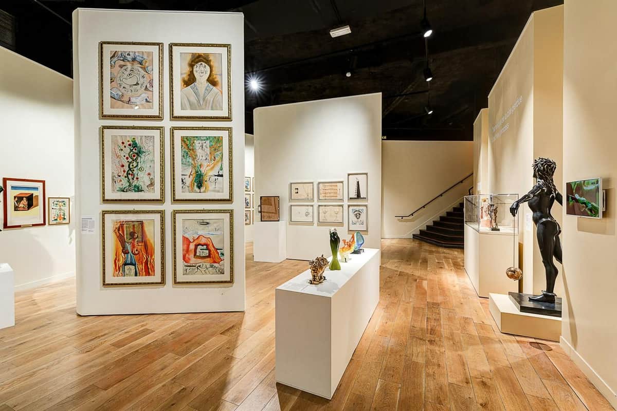 Dalí Paris: Entry Ticket Art gallery featuring framed paintings on walls, sculptures on pedestals, wooden flooring, and a staircase in the background.