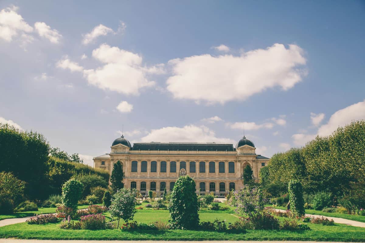 Grande Galerie de l'Évolution: Skip The Line Ticket Large building with domed towers, surrounded by a garden with flowers and shrubs, under a partially cloudy sky.