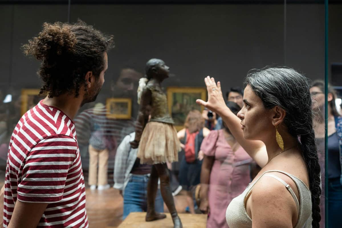 Musée d'Orsay: Skip The Line Ticket + Semi-Private Guided Tour A woman with a braid gestures toward a bronze sculpture of a young girl in a tutu, observed by several people in a museum.