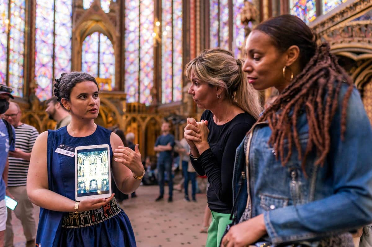 A woman in a blue dress shows an image on a tablet to two others in a Gothic-style building with stained glass windows.
