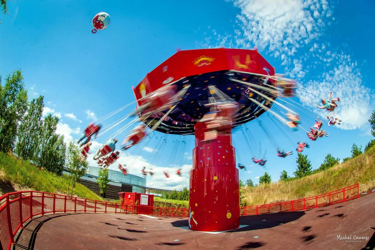 Le Parc du Petit Prince: Fast Track Ticket A red swing carousel in motion against a blue sky with people seated and a balloon floating nearby.