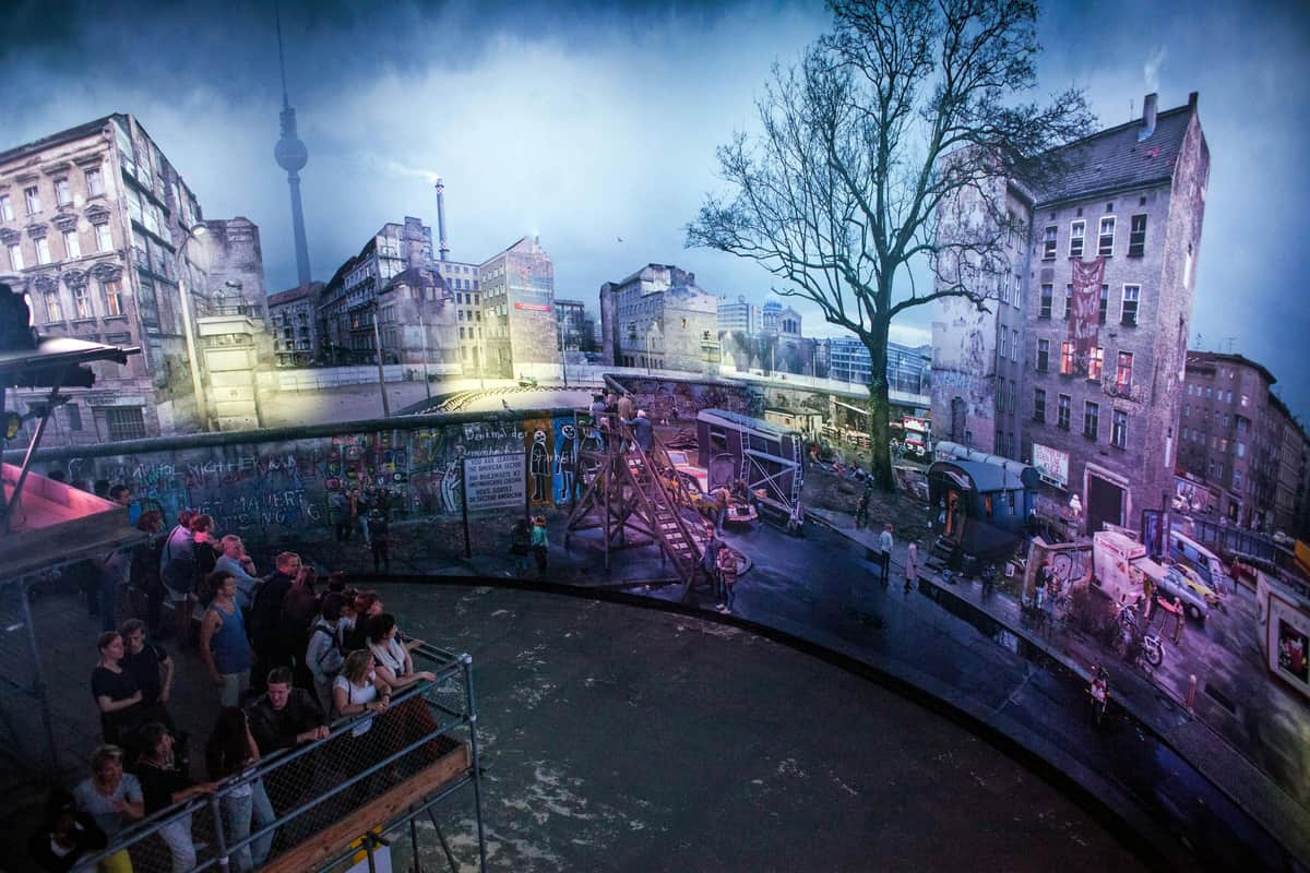 Asisi Panorama - Die Mauer (The Wall): Skip The Line A group of people observe a panoramic display of a cityscape with historical buildings, graffiti, and a large tree.