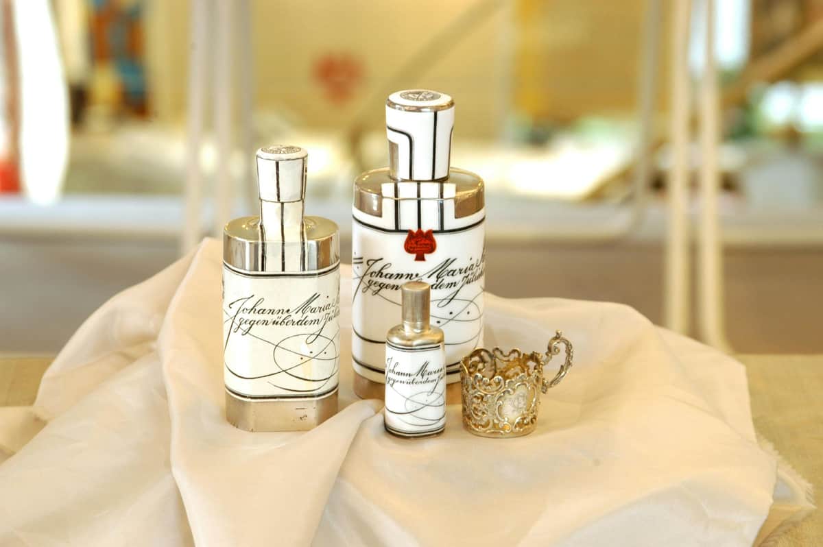 Farina Fragrance Museum: Entry Ticket + Guided Tour Three elegant white perfume bottles with silver caps and decorative script, arranged on a cream-colored fabric next to a detailed metal holder.