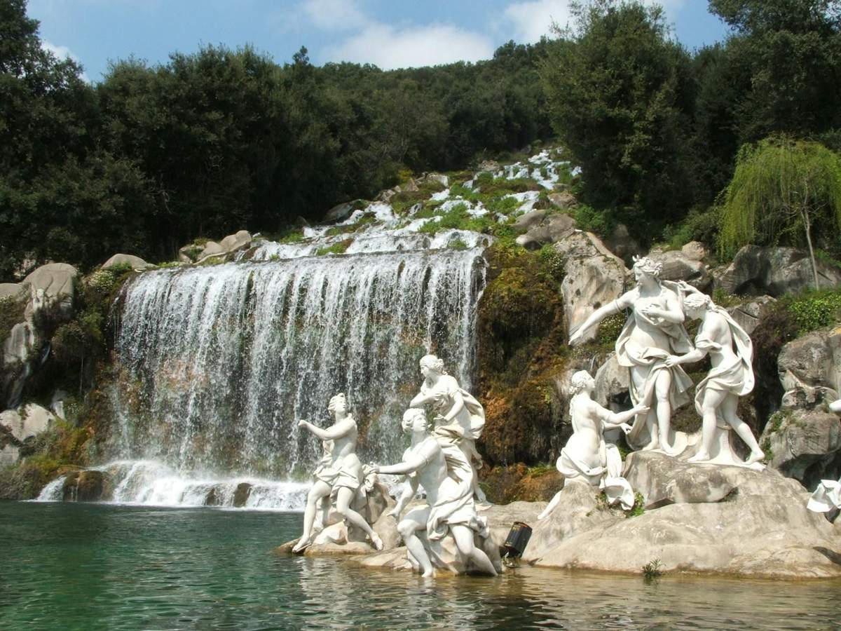 Royal Palace of Caserta: Skip The Line Ticket + Guided Tour White statues depicting human figures stand near a cascading waterfall surrounded by lush greenery.