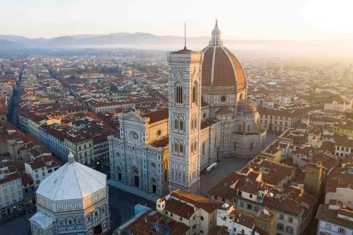 Aerial view of Florence Cathedral with its dome and bell tower, surrounded by city buildings under soft sunlight.
