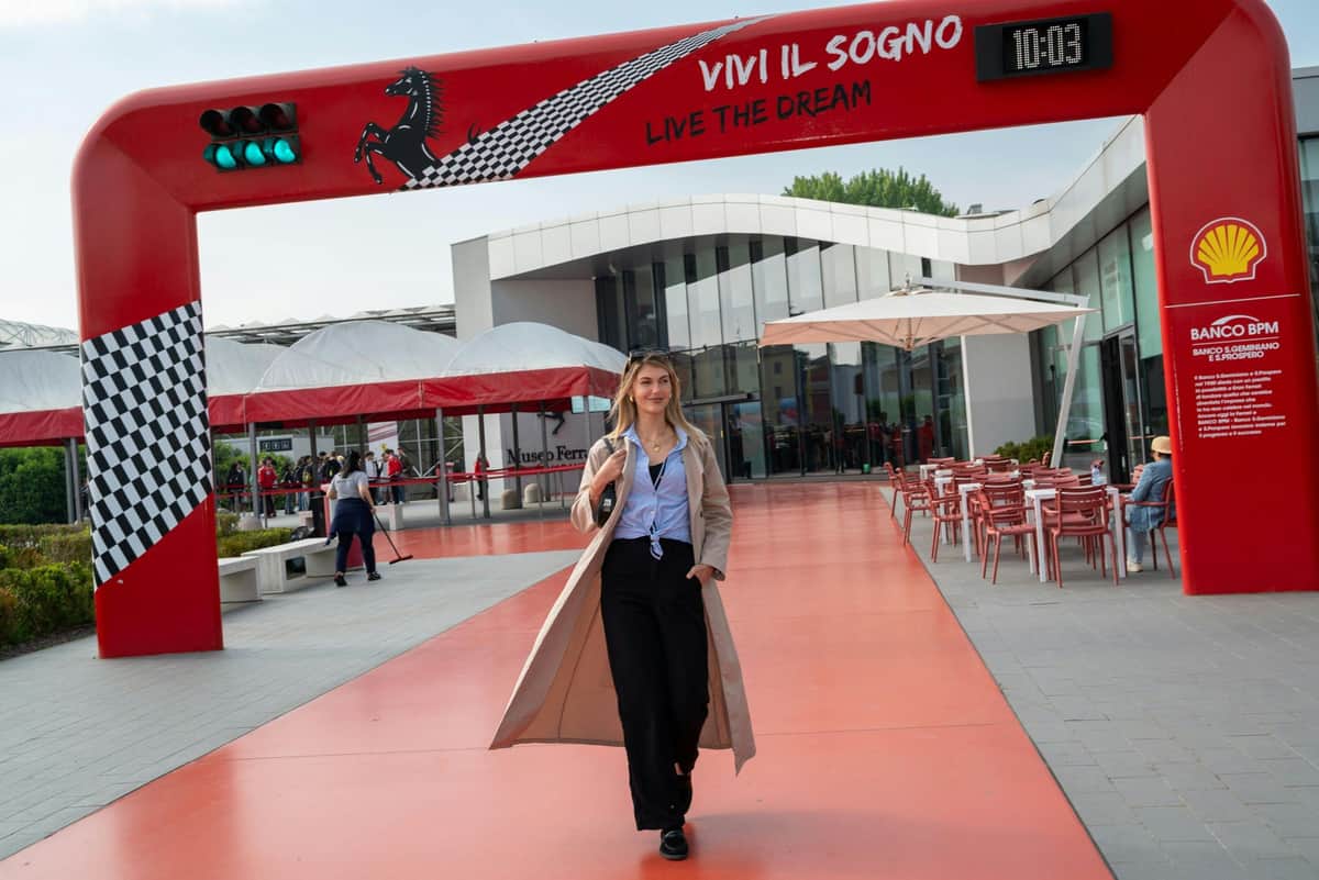 Maranello: Ferrari Museum Skip The Line Entry Ticket + F1 Simulator A woman walks confidently along a red walkway near a modern building, with a Ferrari-themed arch overhead.