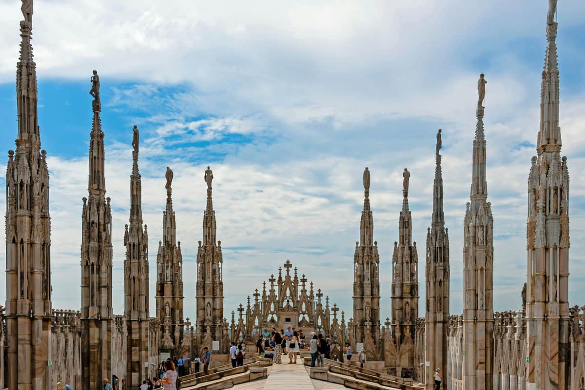 Duomo di Milano: Fast Track Rooftop Access People walking on the roof of Milan Cathedral, surrounded by intricate stone spires and statues under a partly cloudy sky.