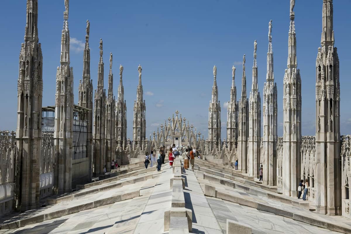 Duomo di Milano: Fast Track Ticket + Rooftop Access People walking on the rooftop of a Gothic cathedral adorned with numerous ornate spires, under a clear blue sky.