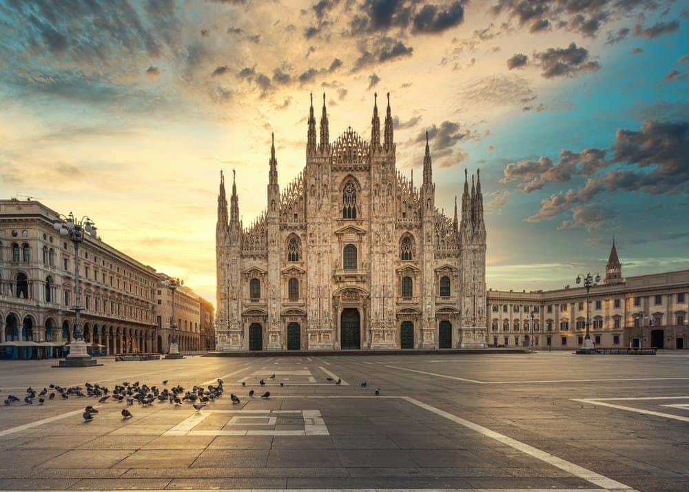 Piazza with scattered pigeons, large ornate cathedral with spires in background, under a dramatic sunset sky with clouds.