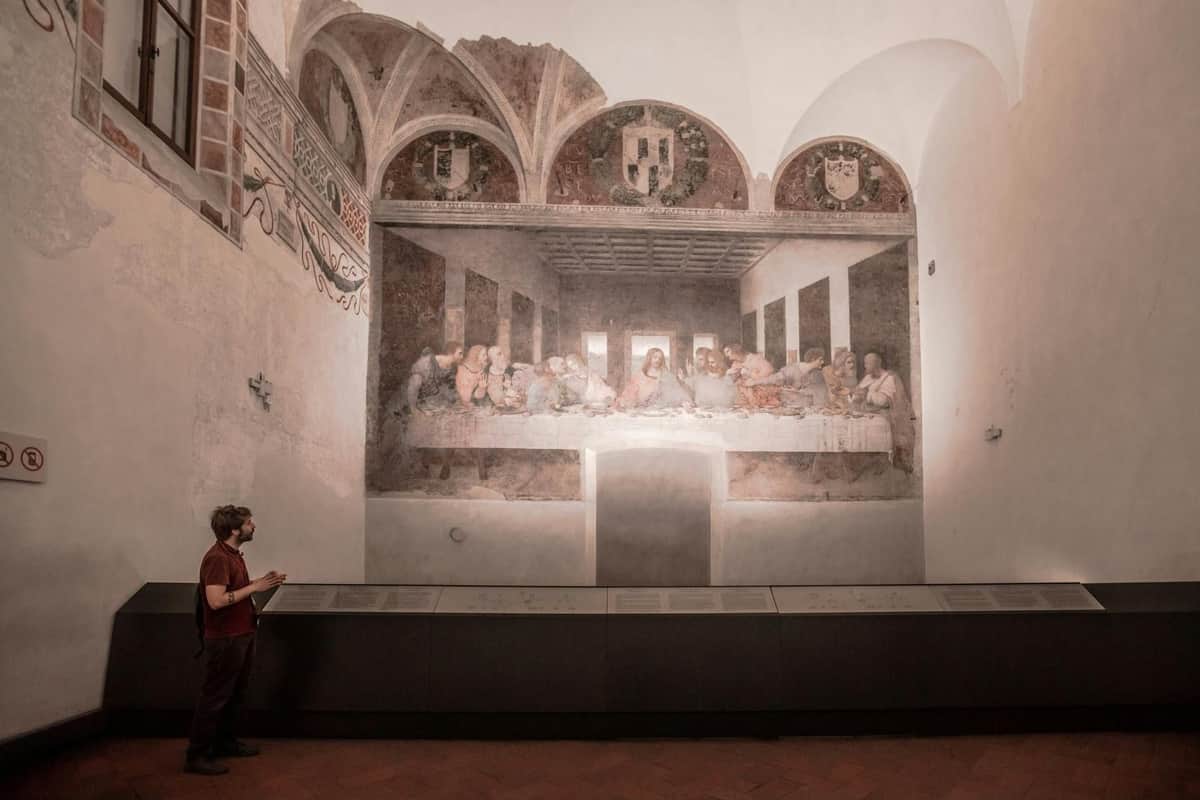 A person stands in front of Leonardo da Vinci's "The Last Supper" mural in a dimly lit gallery.