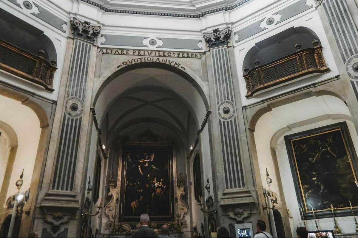 Pio Monte della Misericordia: Skip The Line Ticket + Guided Tour People observing artwork inside an ornate chapel with high arches, columns, and Latin inscriptions on the walls.