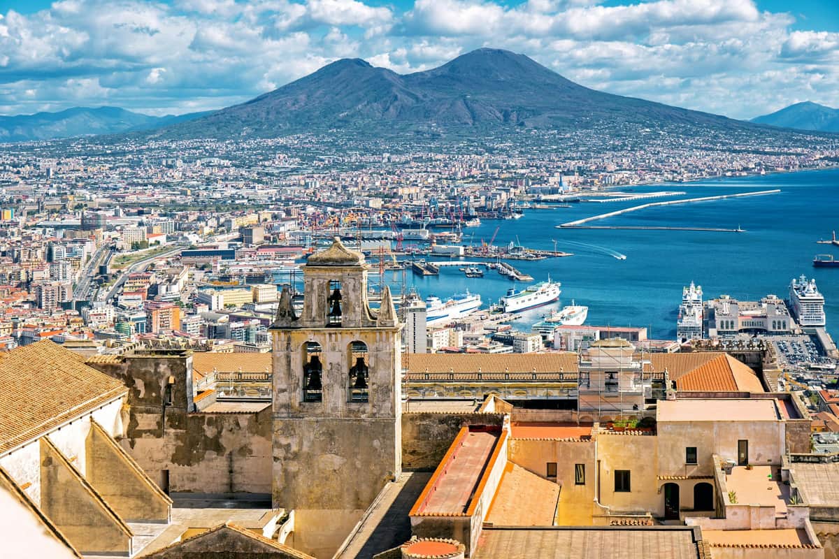 Campania Artecard A cityscape with a historic bell tower in the foreground, a harbor with ships, and a mountain under a blue sky in the distance.