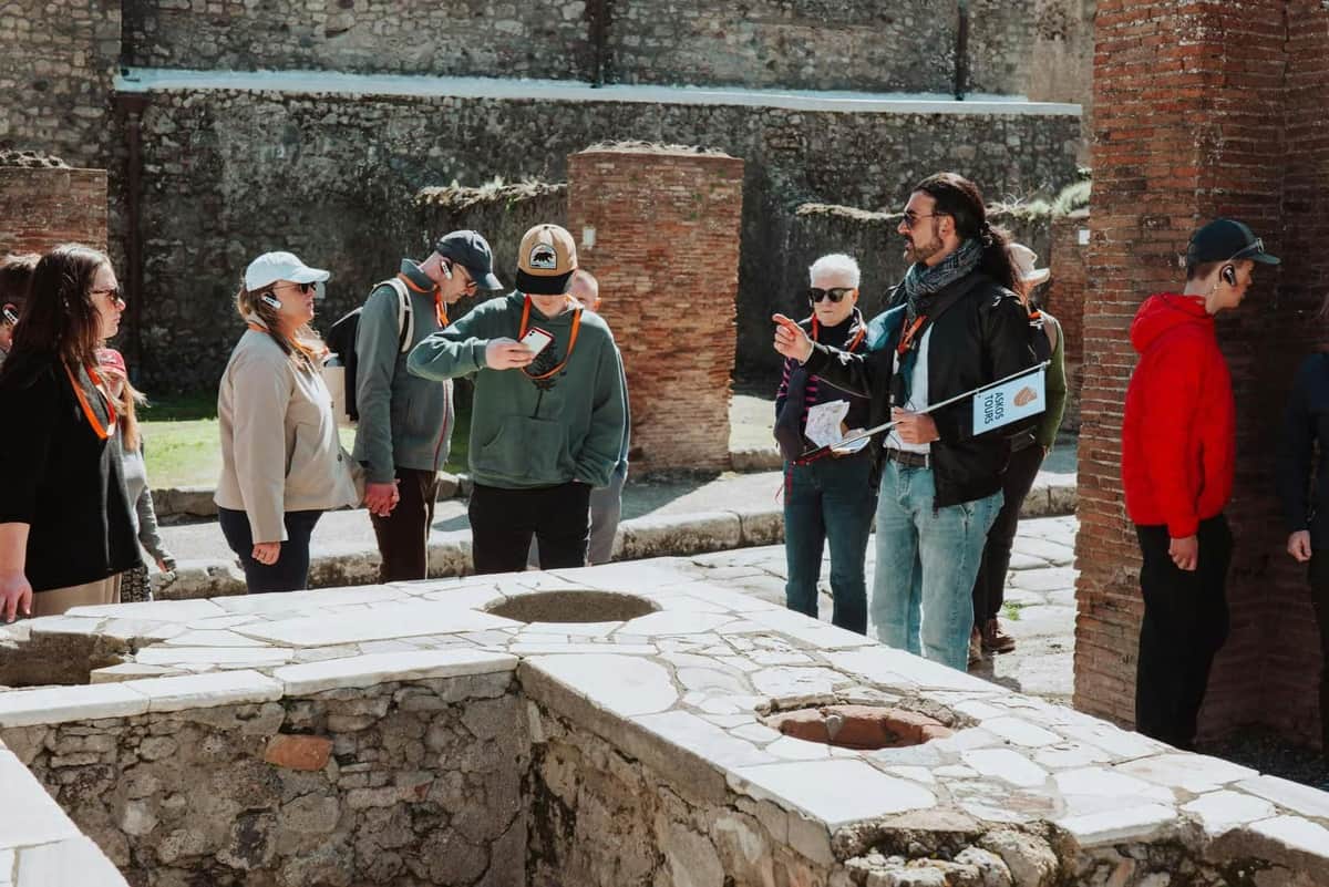 Um grupo de pessoas com cordões laranja encontra-se junto a ruínas antigas, tendo como pano de fundo paredes de pedra e áreas relvadas.