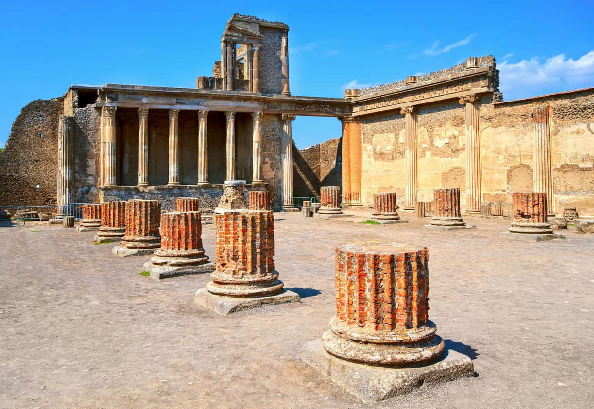 Pompeii: Guided Tour Ancient ruins with multiple broken columns and a large stone building featuring tall pillars and partially intact walls under a blue sky.