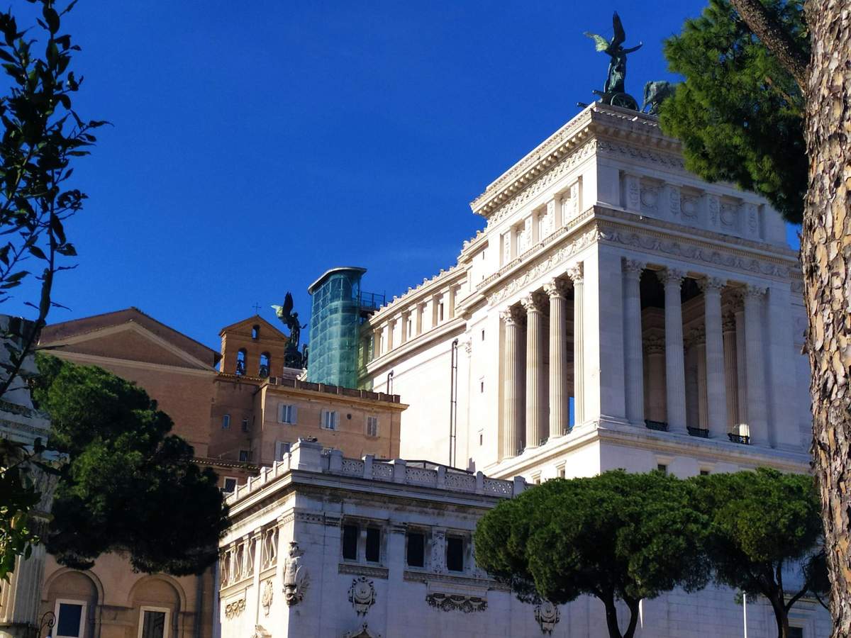 Historic buildings with columns and statues against a blue sky, partially obscured by trees in the foreground.