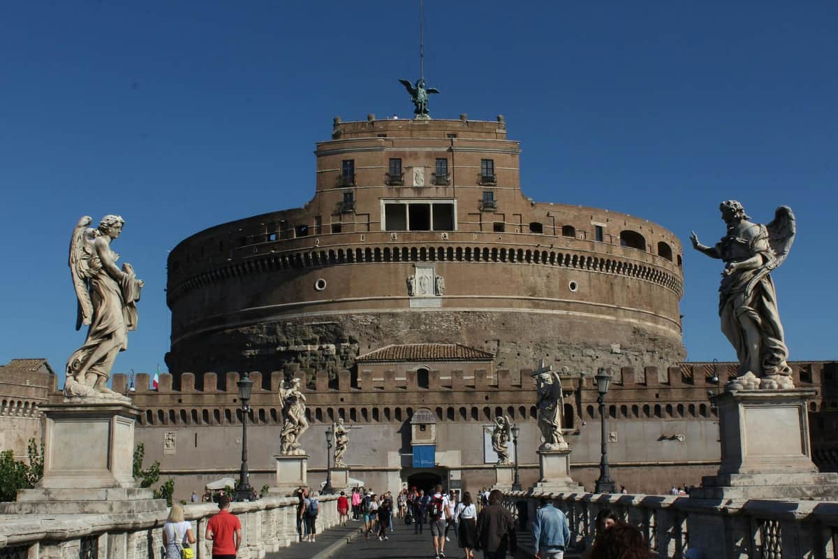 Large, circular brick fortress with a statue on top, viewed from a crowded pedestrian bridge adorned with statues under a clear blue sky.