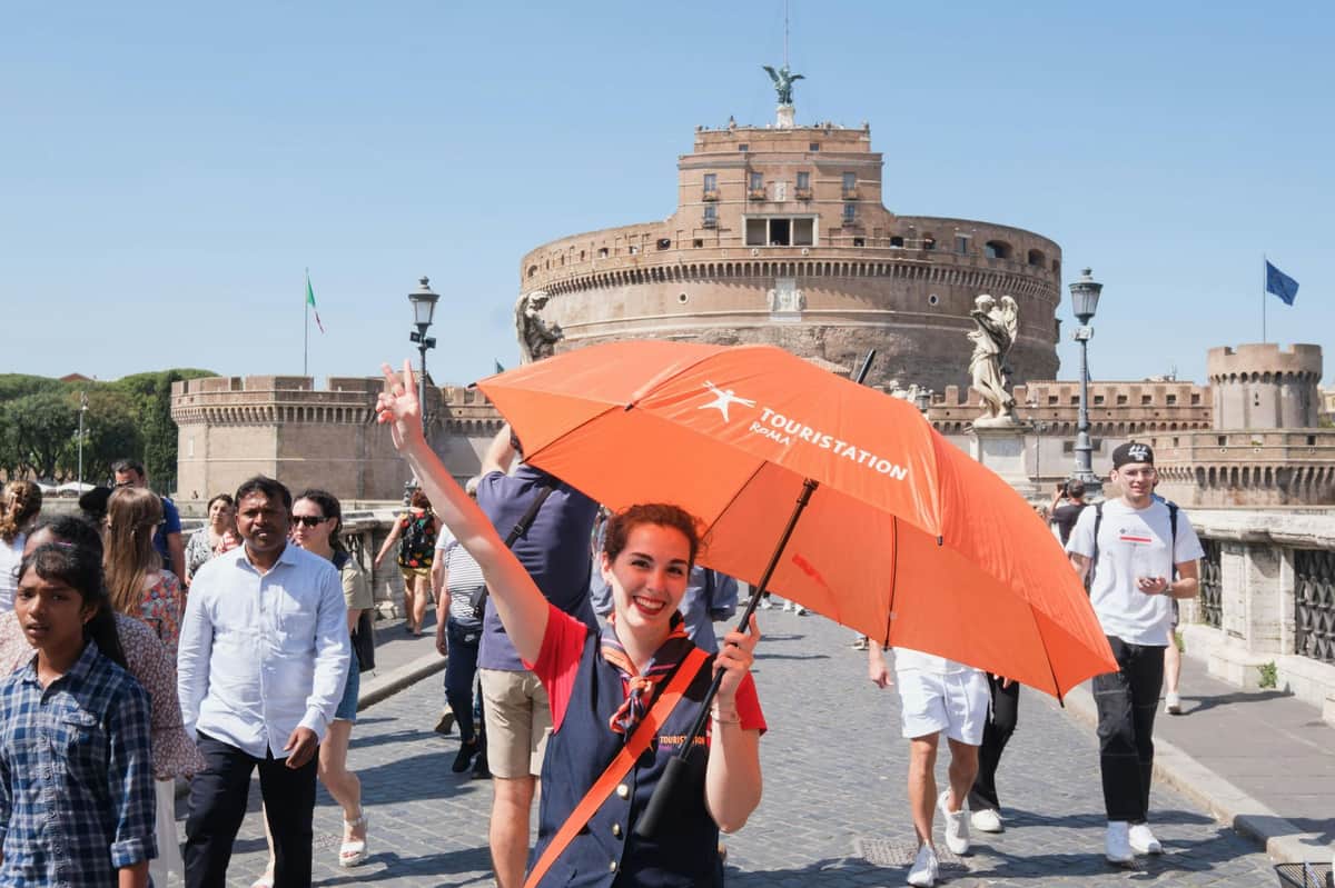 Castel Sant'Angelo: Skip The Line Ticket + Multimedia Video A cheerful woman holding an orange umbrella stands on a bridge with the Castel Sant'Angelo and tourists in the background.