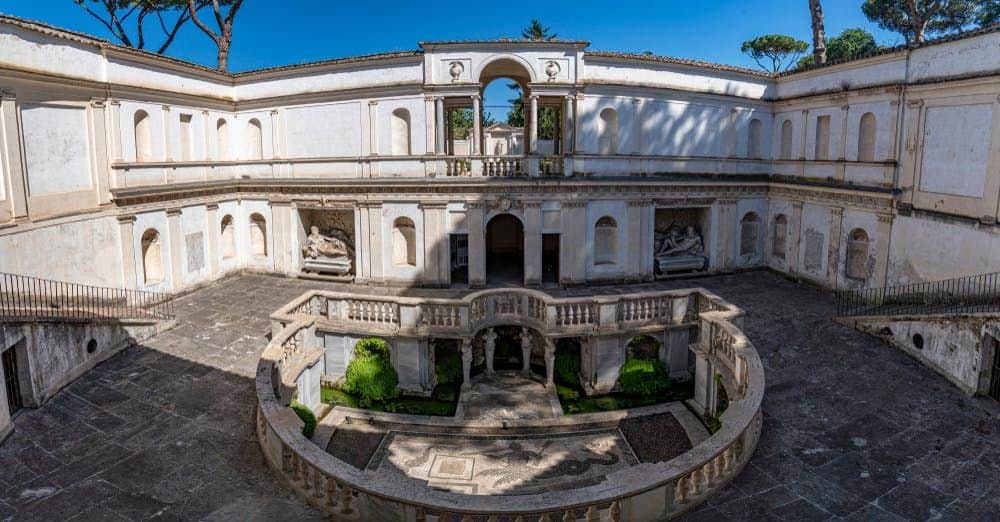 Stone courtyard with intricate balustrades, archways, and statues, surrounded by a two-story building under a clear blue sky.
