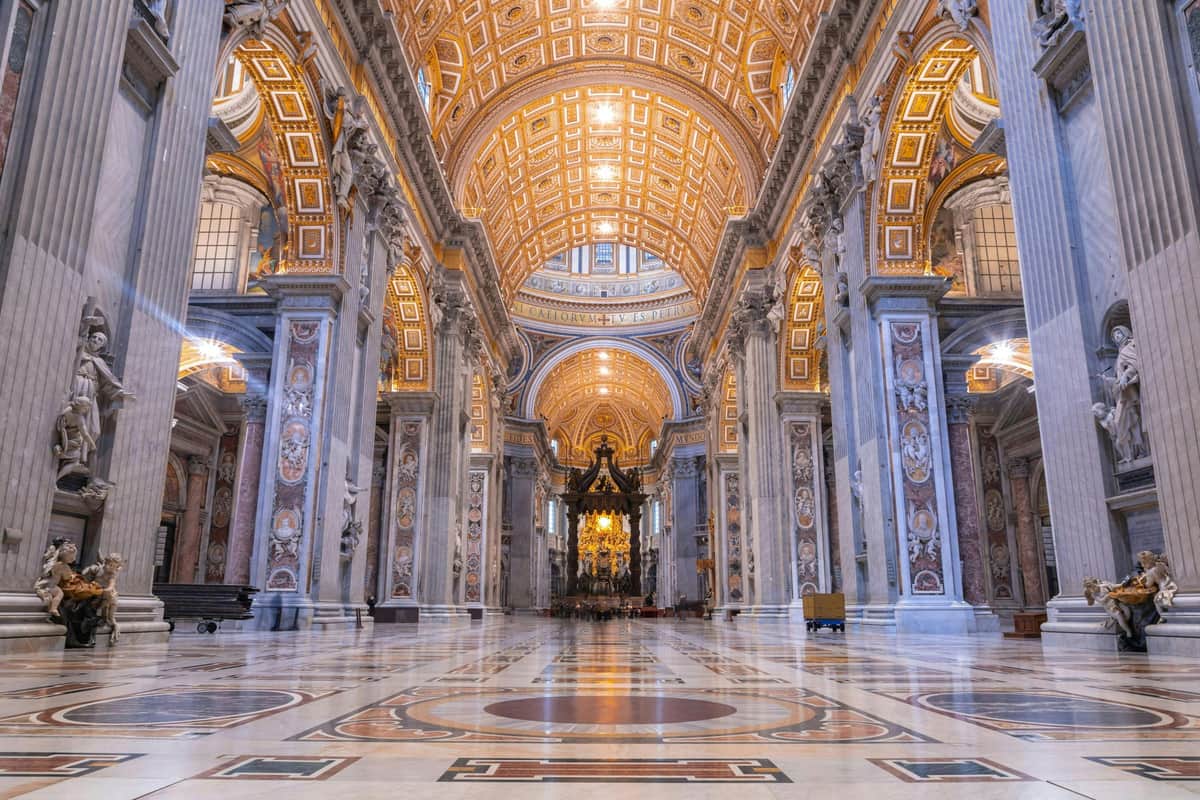 Basílica de São Pedro: Entrada rápida + audioguia Interior ornamentado de uma grande catedral com um teto decorado, colunas de mármore, arcos, padrões intrincados no chão e um altar ao longe.