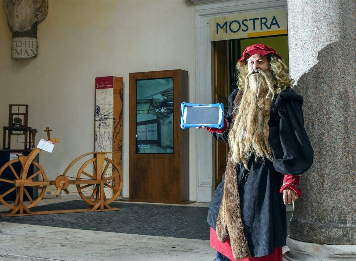 Treasure Hunt with Leonardo da Vinci A person in historical costume with a long wig and beard holds a blue tablet while standing near a building entrance labeled "MOSTRA."