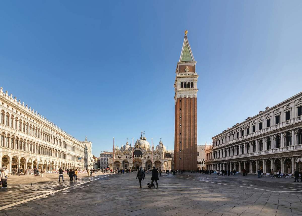 Doge's Palace & St. Mark's Basilica: Guided Tour A wide plaza with a tall brick bell tower, a large ornately designed church, and people walking around on a clear day.