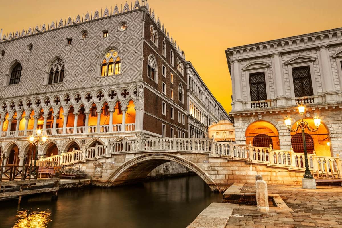 St. Mark's Square Prison Palace: Entry Ticket Historic buildings with ornate facades and arches are connected by a bridge over a canal at sunset.