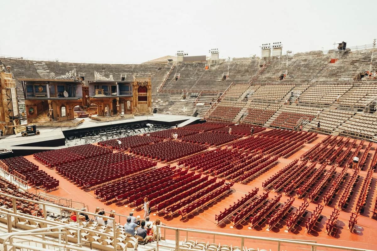 Verona Arena: Fast Track Ticket A large, empty amphitheater with rows of red chairs facing an elaborate stage. Few people are scattered in the seating area.