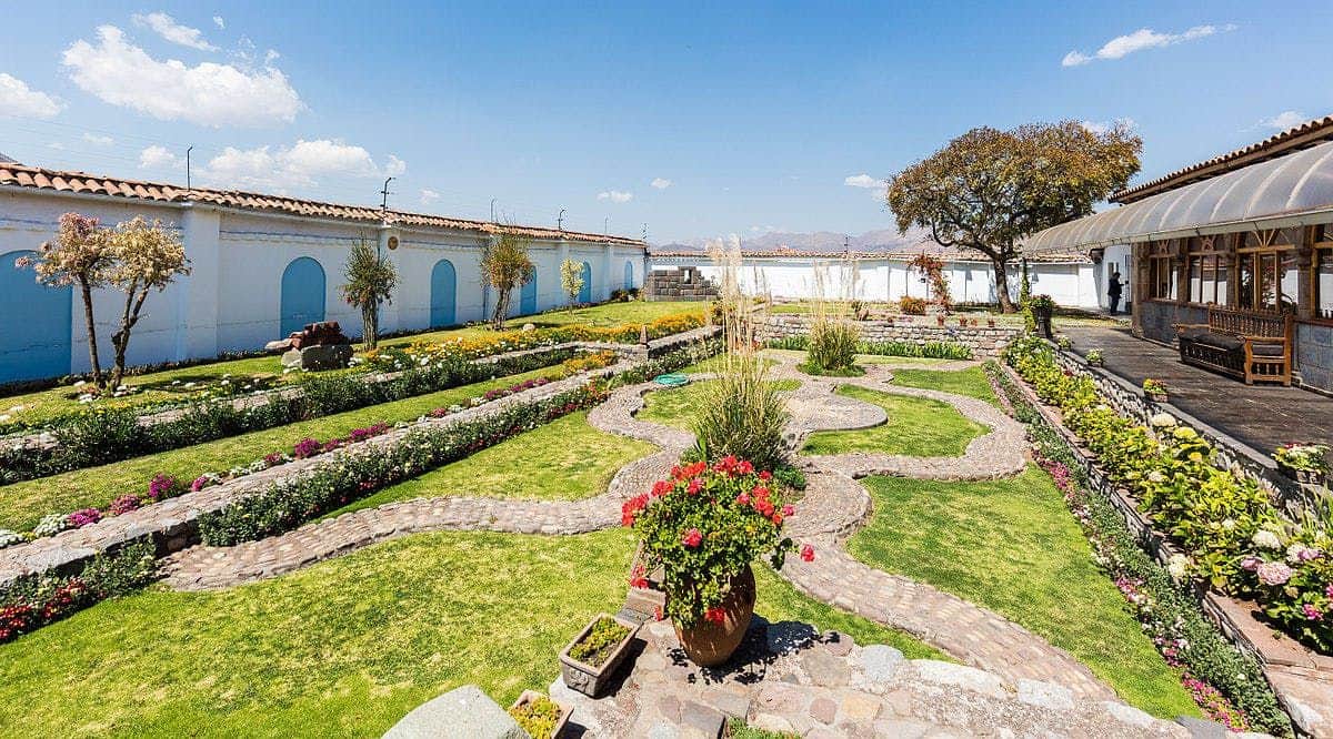 Lush garden with stone pathways, colorful flowers, and potted plants, bordered by a white building with blue doors under a clear sky.