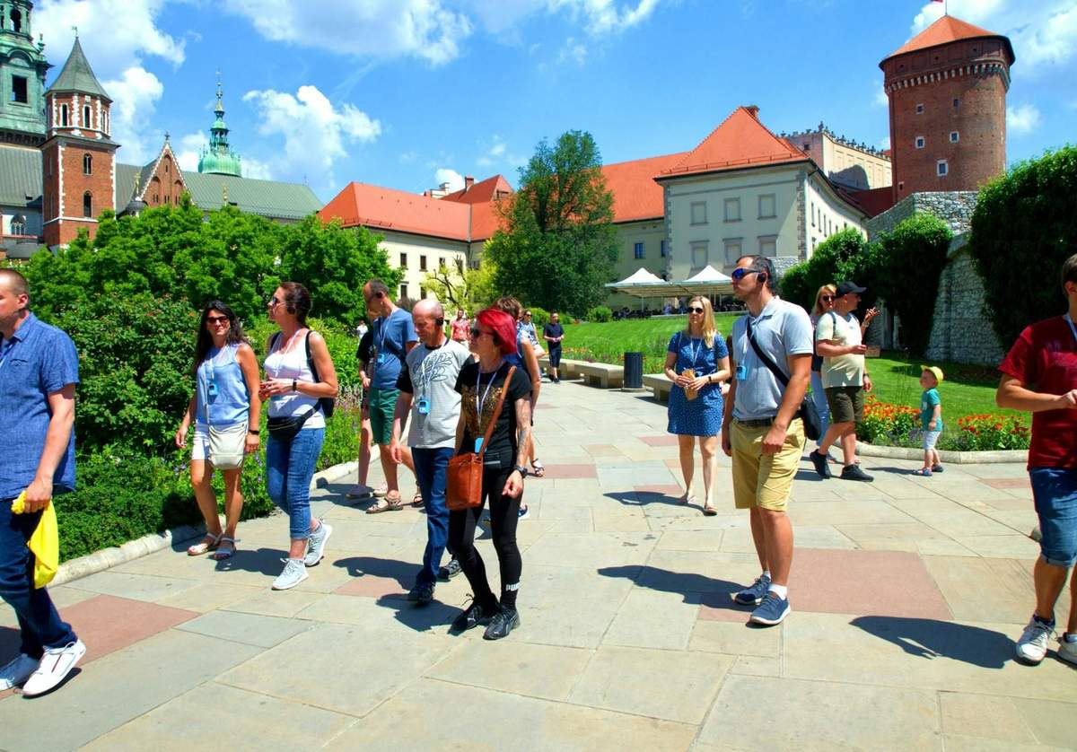 Wawel Castle Guided Interior Tour: Skip-the-Line A group of people walks on a paved path with historic buildings, greenery, and a clear blue sky in the background.