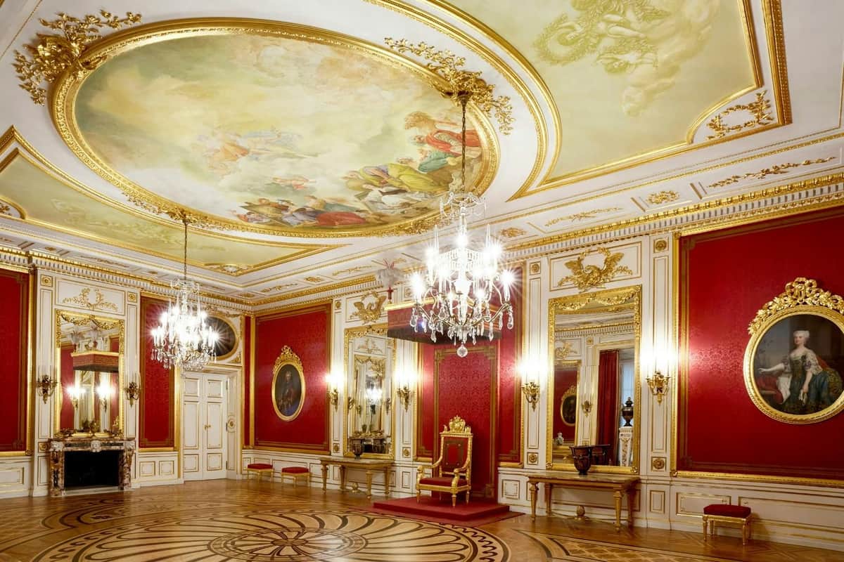 An ornate room with red walls, chandeliers, a painted ceiling, and a central throne on a red carpet.