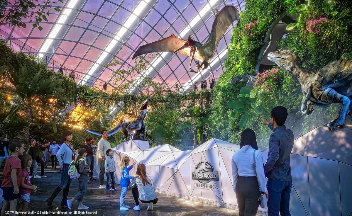Gardens by the Bay: Flower Dome & Cloud Forest Entry Ticket People observe animatronic dinosaurs in a futuristic, glass-ceilinged exhibit with lush plants and the "Jurassic World" logo.